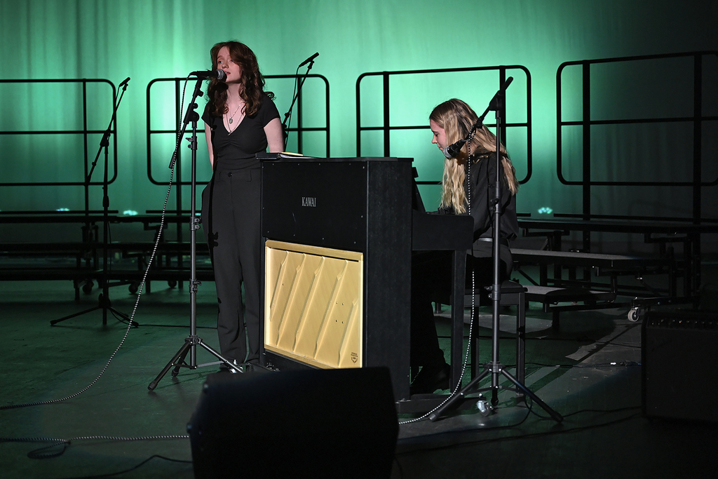 Two women perform on stage with microphones, one standing and singing, the other seated at a piano.