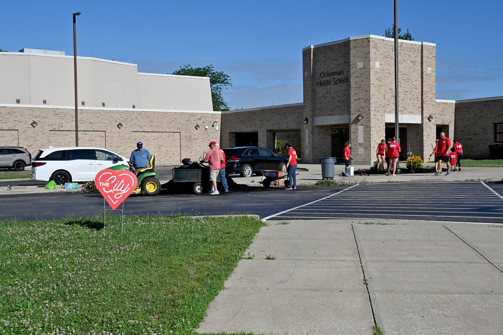 A building with people in red shirts, a lawn mower, and a heart sign in a grassy area.