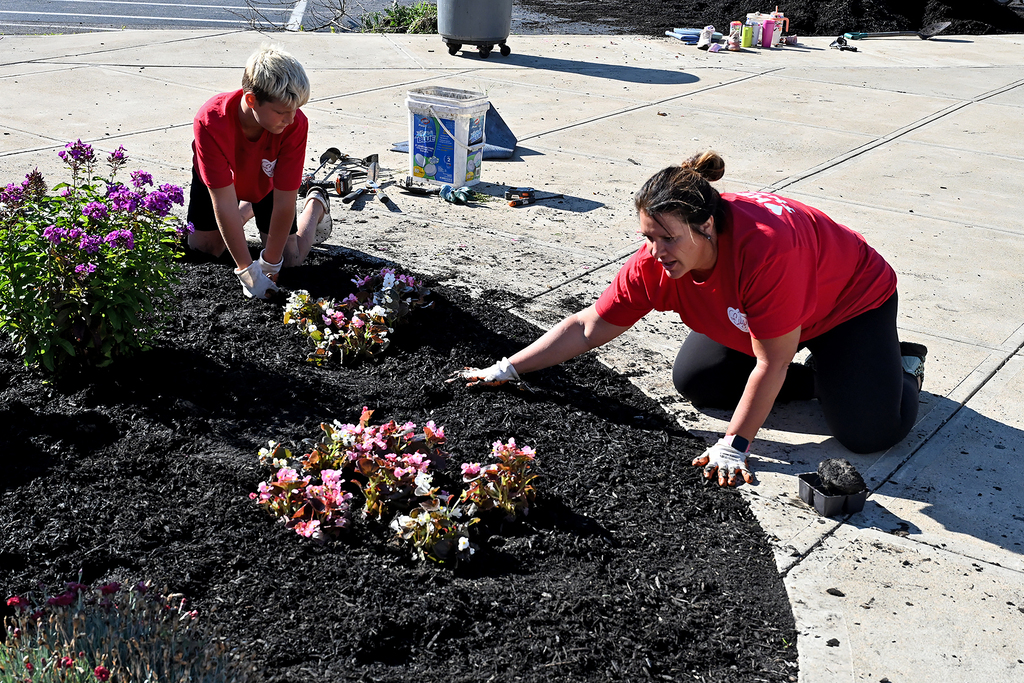 Two people in red shirts plant flowers in a garden bed on a sidewalk.