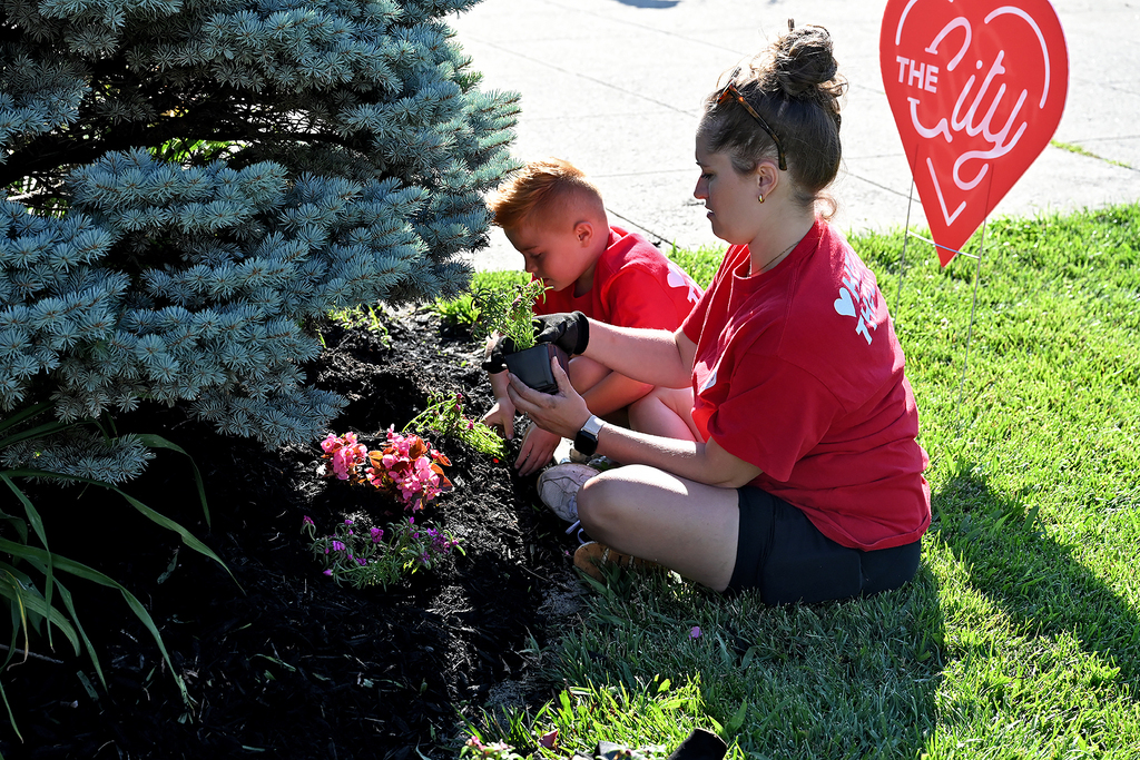 A woman and child planting flowers in a yard with a red heart sign in the background.