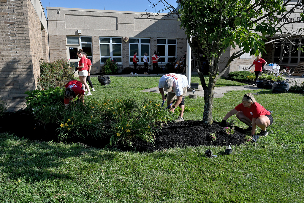 People in red shirts and shorts work in a garden with flowers and a tree. A building is in the background.