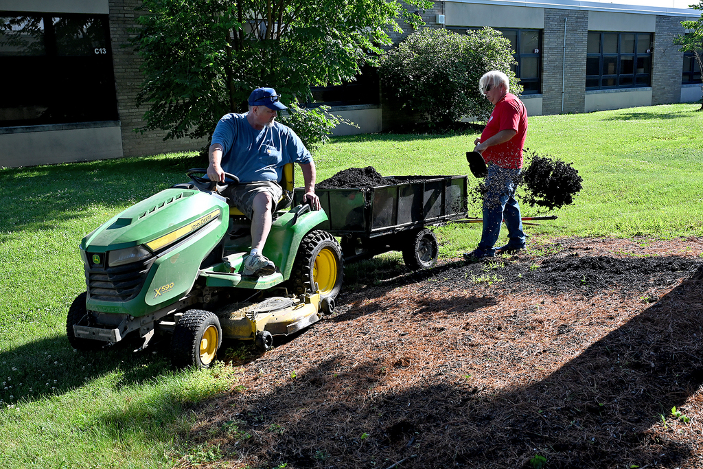 Two people outside, one on a green lawnmower, another holding plants. A building with glass windows in the background.
