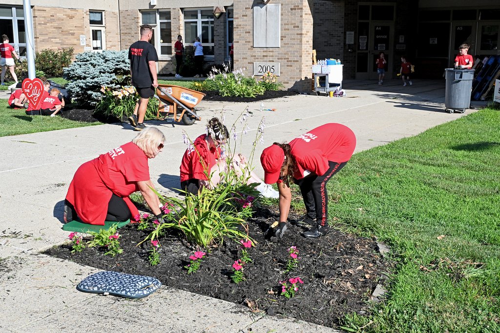 Four people in red shirts and caps are planting flowers in a garden. Two people are bending down, and two are standing.