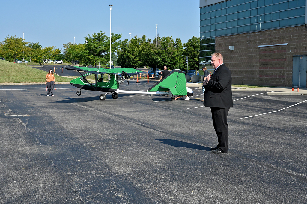 A man in a suit stands in a parking lot with a green airplane behind him.