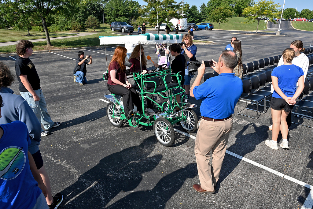Several people observe a person seated on a green four-wheeled bike in a parking lot. One person holds a camera.