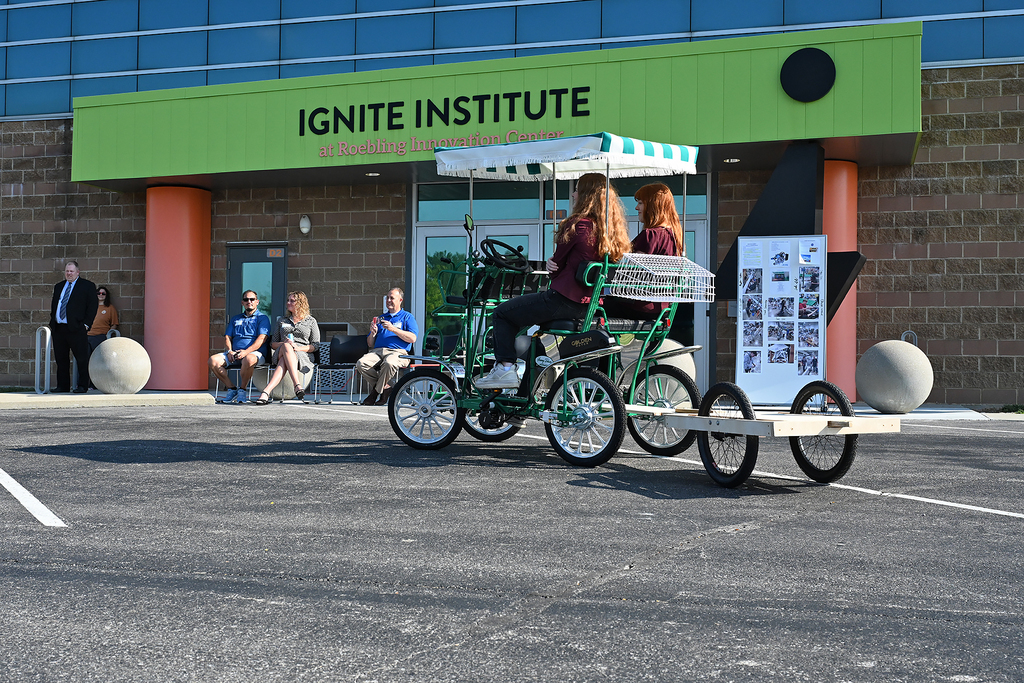 Two women riding a four-wheeled bike in front of the Ignite Institute building. People sit on the sidewalk.