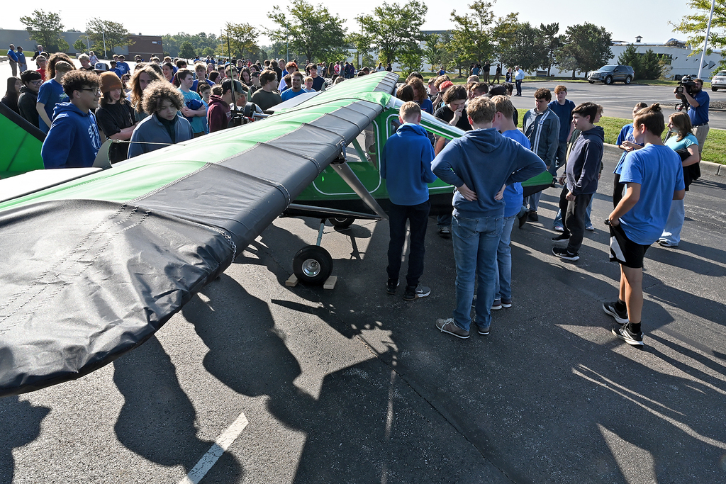 Many people gather around a green and black airplane in a parking lot. Some wear light blue shirts.