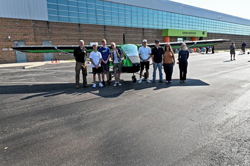 A group of people stands in front of a small plane on a paved surface. The building behind them has glass windows.