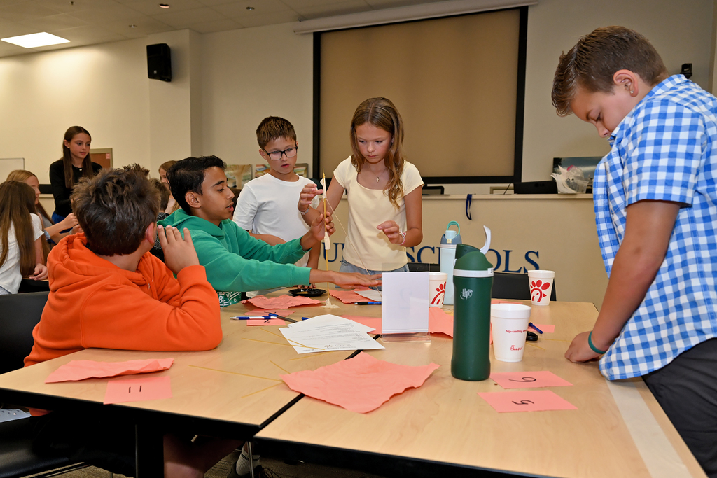 Group of children at a desk, engaged in an activity, with papers, markers, and a water bottle.