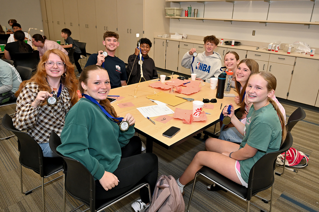 A group of young adults wearing medals sit around a table in a classroom, smiling and posing.