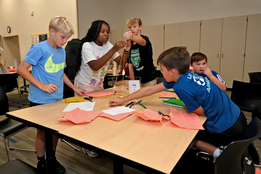 Group of children at a table in a classroom, working with colored paper, scissors, and markers.