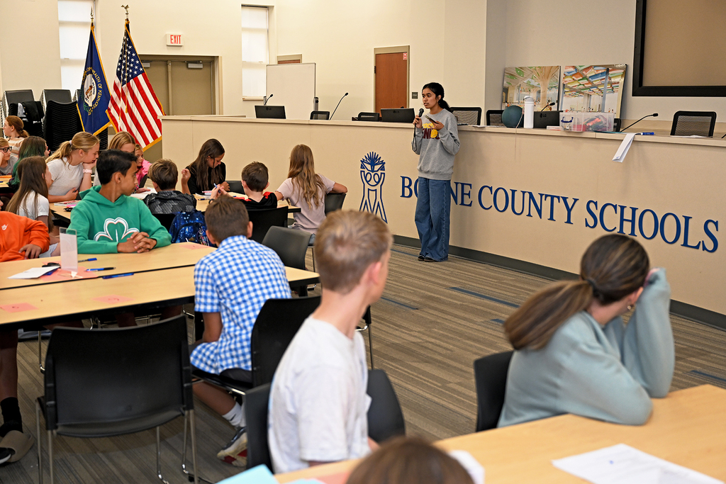 A classroom with students seated at tables, one standing at a podium, an American flag, and a wall that reads "BONE COUNTY SCHOOLS".