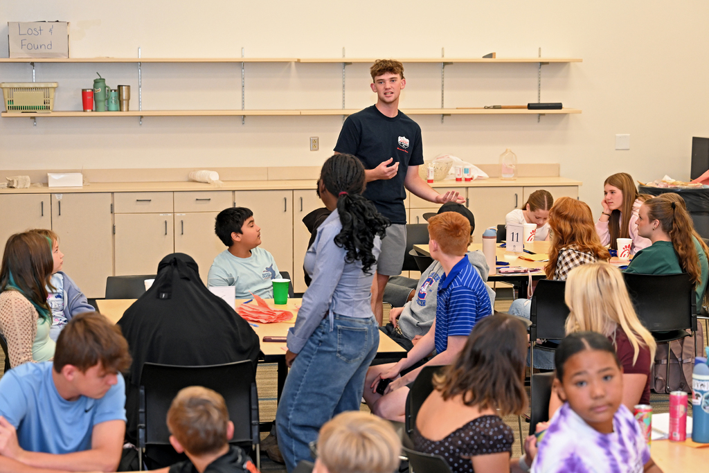 A man speaks to a group of students seated at tables in a classroom. Shelves line the wall behind him.