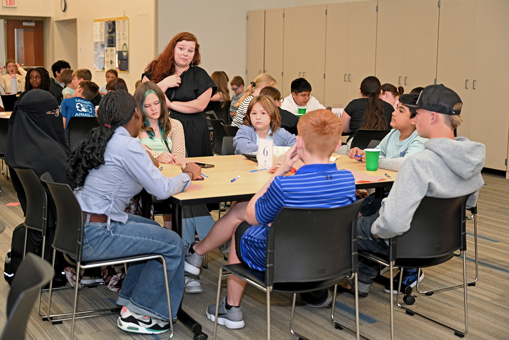 A group of students and an adult sit around a table in a classroom. One adult stands.
