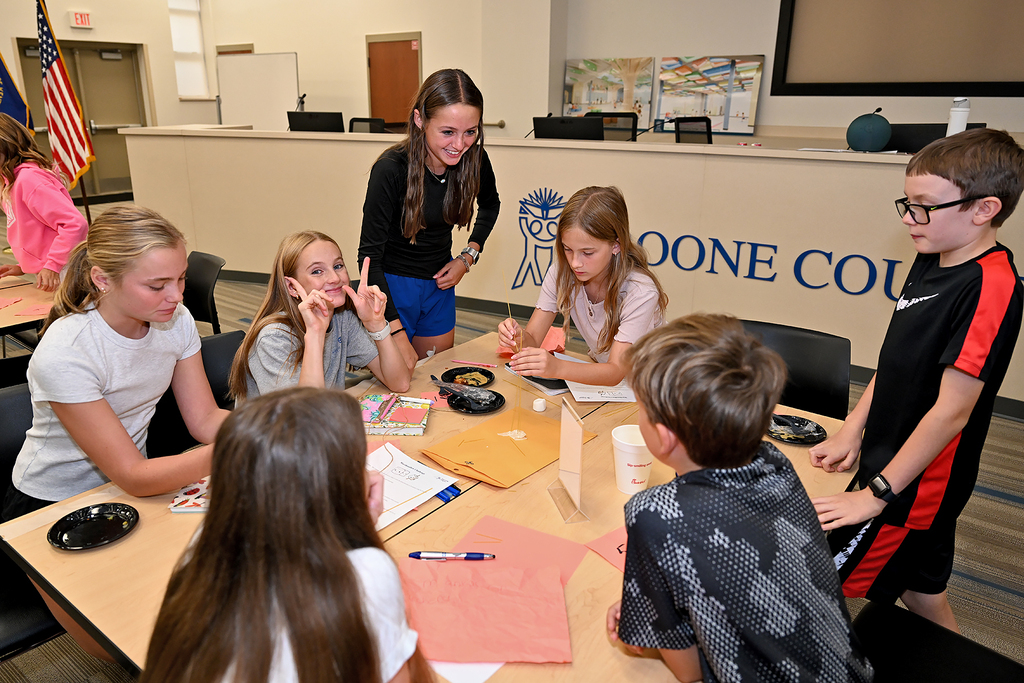 Several children seated around a table, looking at paper and writing materials, one girl stands and gestures.