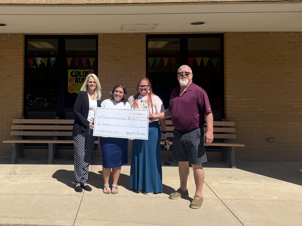 Four adults stand in front of a brick building. They hold a large check. Two women wear skirts, one holds a sign.