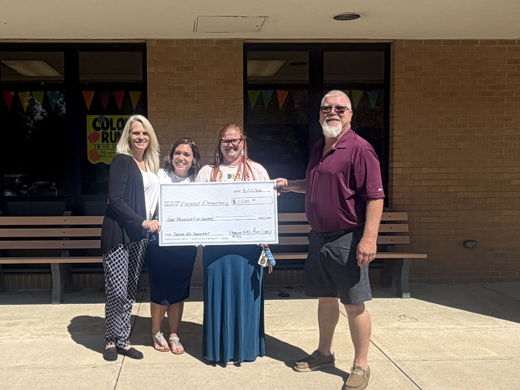 Four individuals stand in front of a building, holding a large check. The woman on the left wears sandals and a skirt.