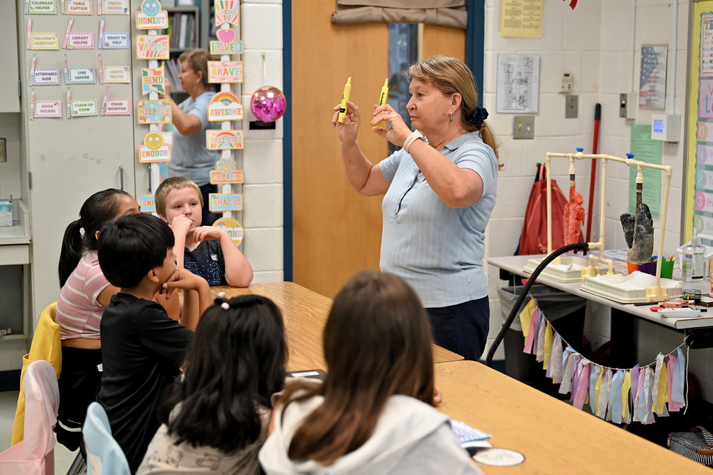 A woman holds yellow objects in a classroom while several students sit in chairs, watching.