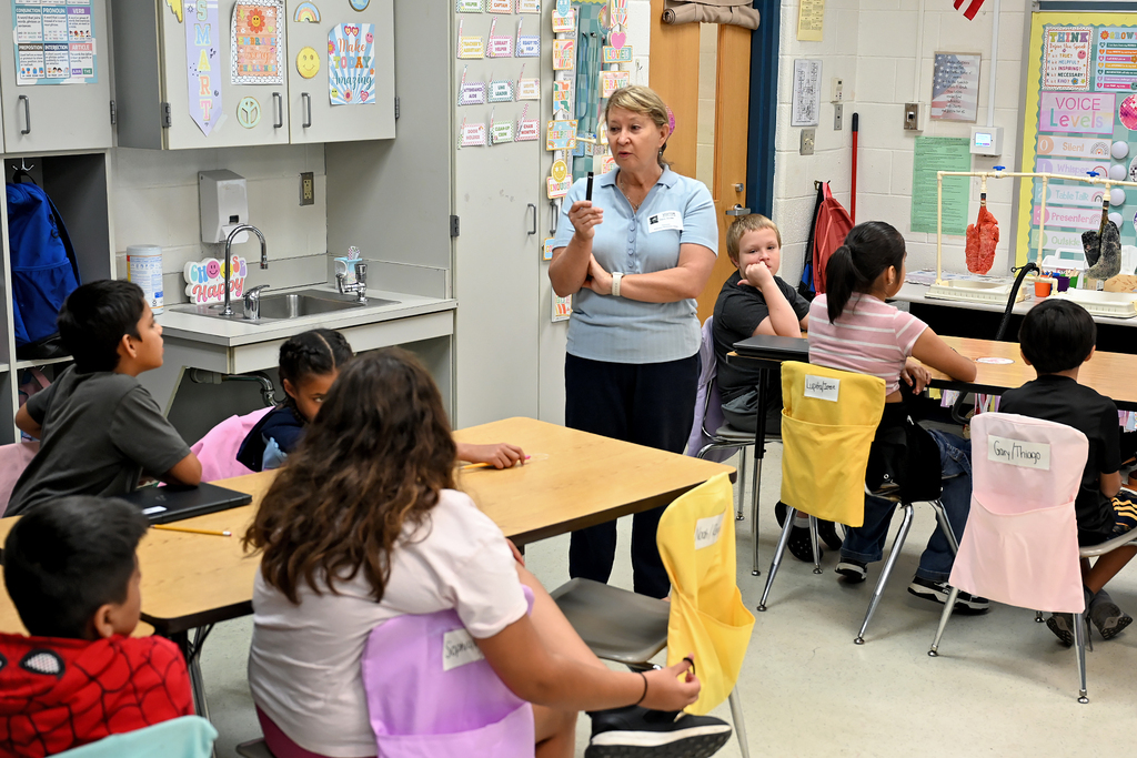 An adult stands at a desk in a classroom, addressing a group of students seated at their desks.