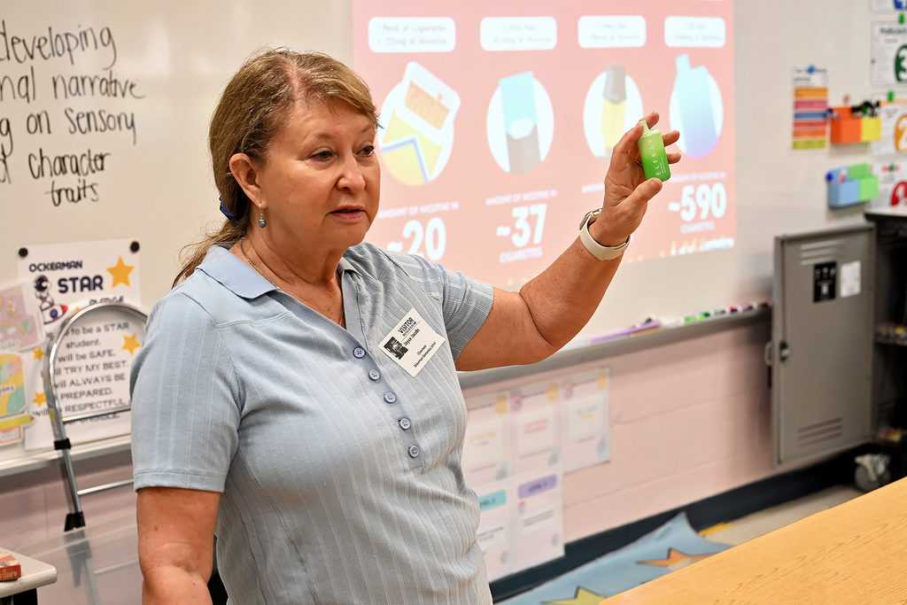 A woman in a blue shirt stands in a classroom, holding a green object, and teaching near a whiteboard.