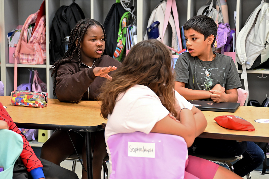 Four children at a table. Three with faces visible. One girl speaking with hands. 