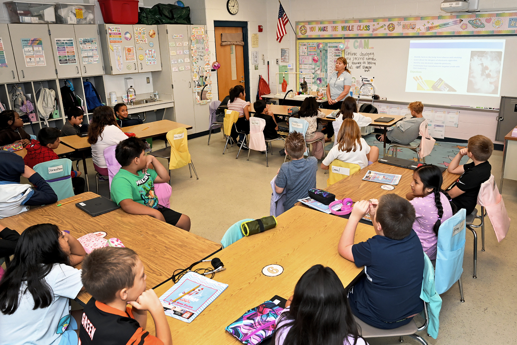 A classroom with children seated at tables, an adult standing by a projector screen on the wall.