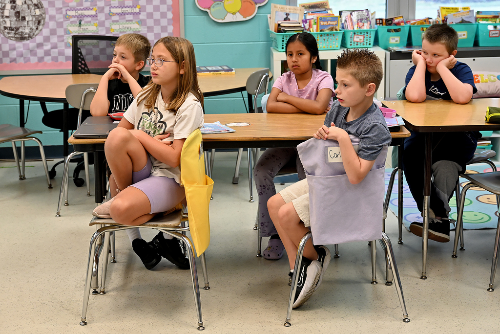 Five children in a classroom setting with arms crossed, seated around tables with books and materials.