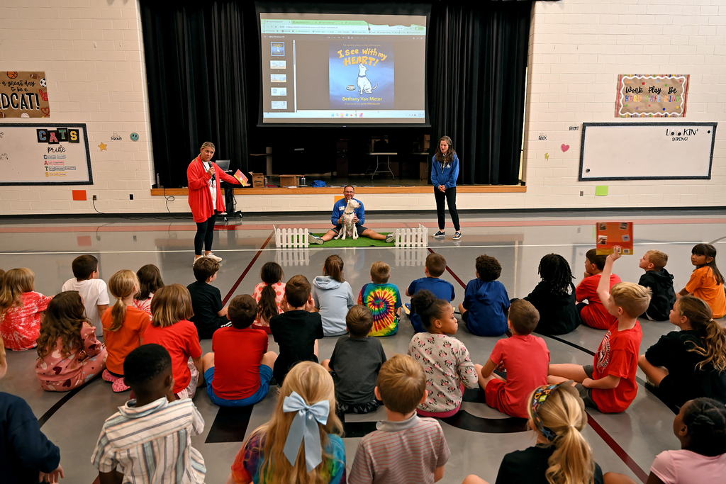 Children and two adults sit in a circle on a floor. A screen displays an image on the wall behind them.