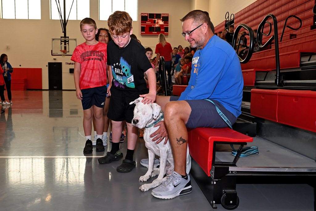 A man and two children interact with a dog in a gym. The man is seated on a bench while the dog rests on his lap.