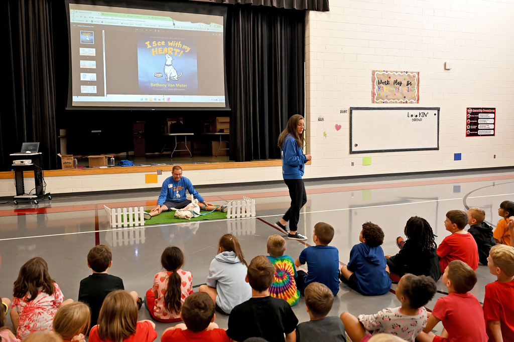 A woman stands by a projector screen showing a dog. Children sit on the floor watching, some wearing backpacks.
