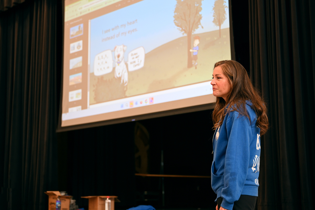 A woman stands in front of a projector screen, showing an animated presentation. She wears a blue sweatshirt.