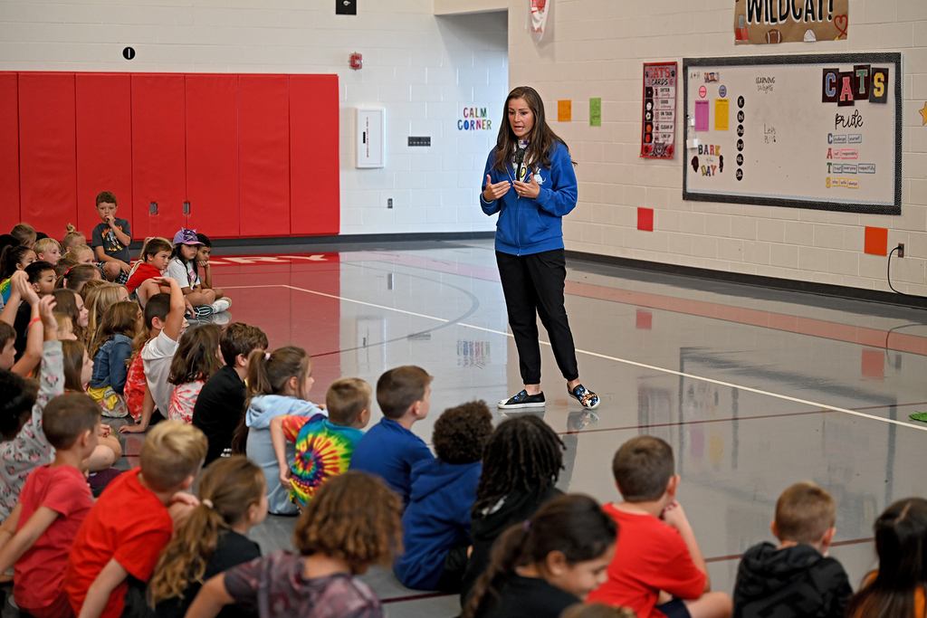 A person stands in front of a seated group in a gym, addressing the children.