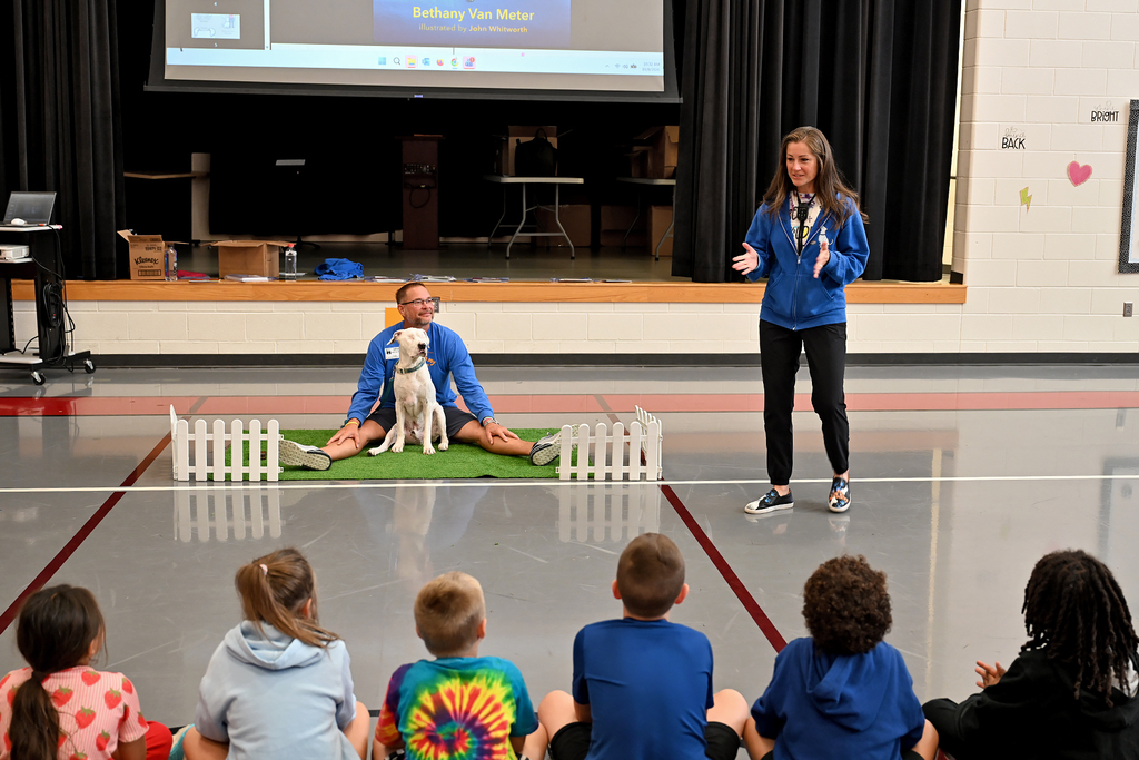 A woman in blue and black stands with a dog on a mat; a man in blue sits on the mat. Kids in hoodies and T-shirts sit on the floor watching.