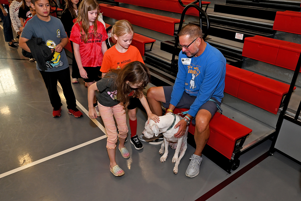 Man in blue shirt and sunglasses sits on bleachers, petting a white dog while several children watch.