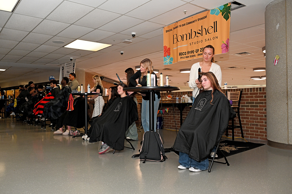 A group of students sitting in chairs with capes while a woman uses a hair dryer.
