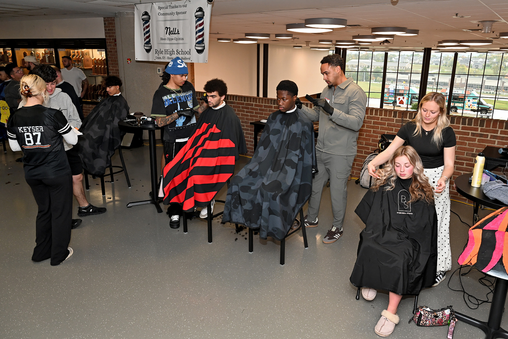Several people in a room with brick walls, receiving haircuts from staff members. Tables and chairs are set up in the area.