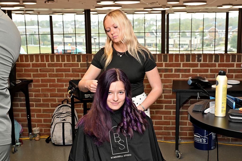 A woman with purple hair in a salon chair gets her hair done by another woman.