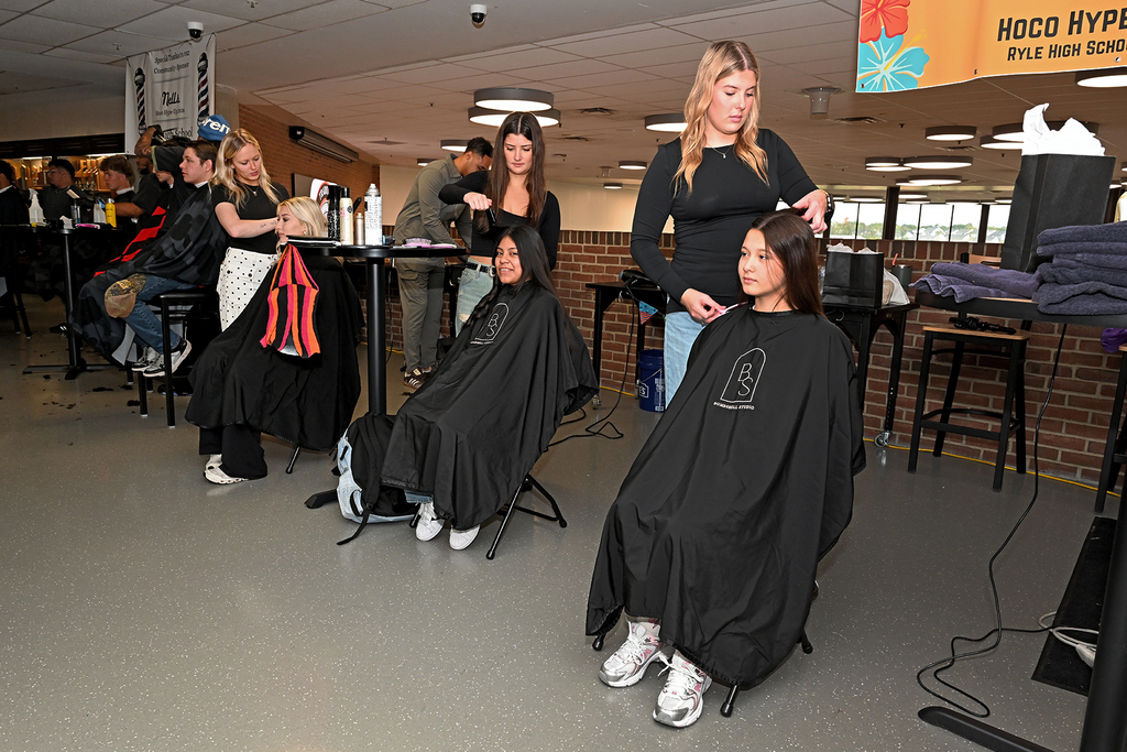 Several students getting their hair cut in a school gym. People seated and standing around.