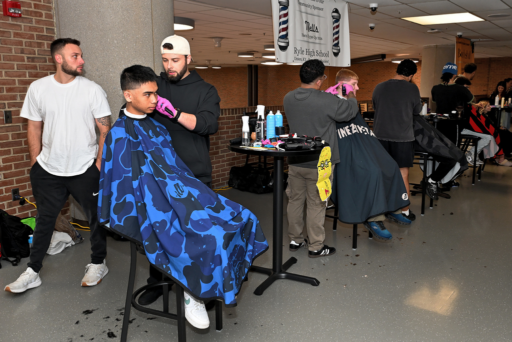 People getting haircuts in a busy room with brick walls and white ceiling lights.
