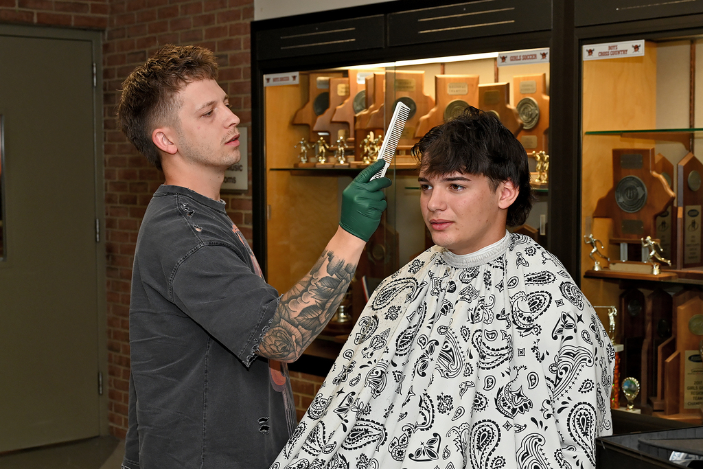 Man in green gloves cutting a boy's hair; he wears a patterned cape. Behind them, a glass case holds sports trophies.