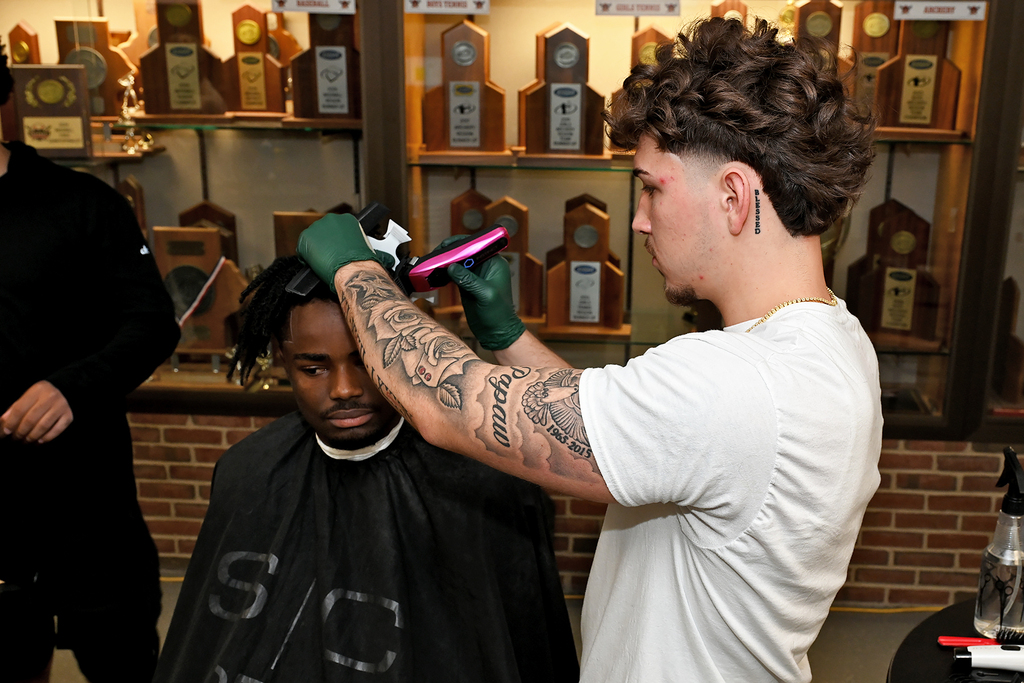 Barber giving a haircut to a seated man. Behind them are shelves with trophy displays.