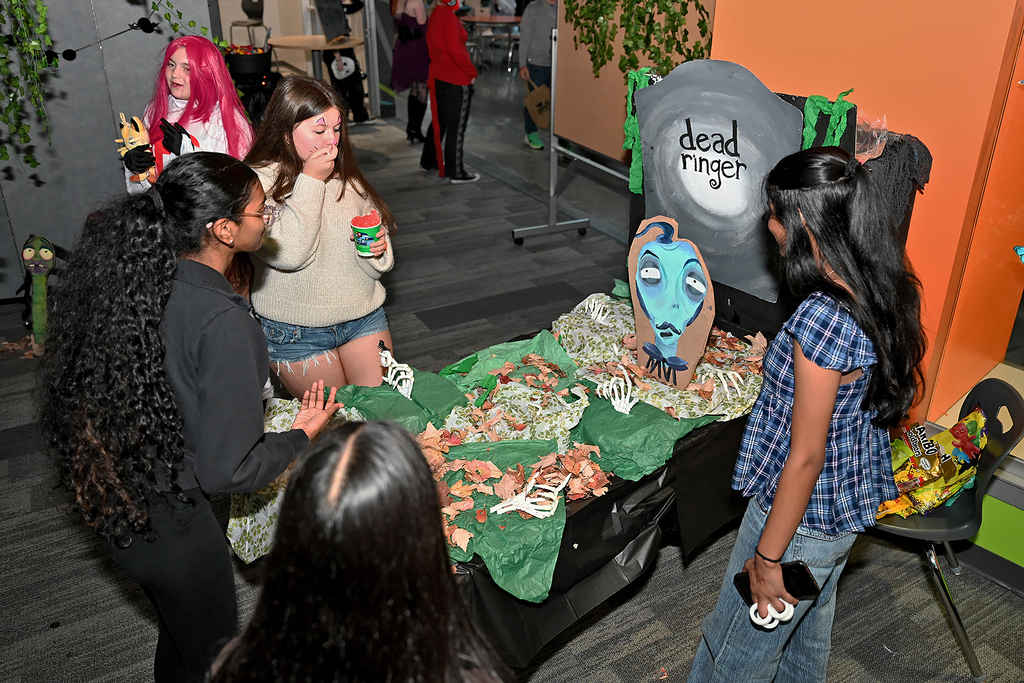 Several students gathered around a table with a green cloth, bones, and a blue face. Two girls are standing behind the table.