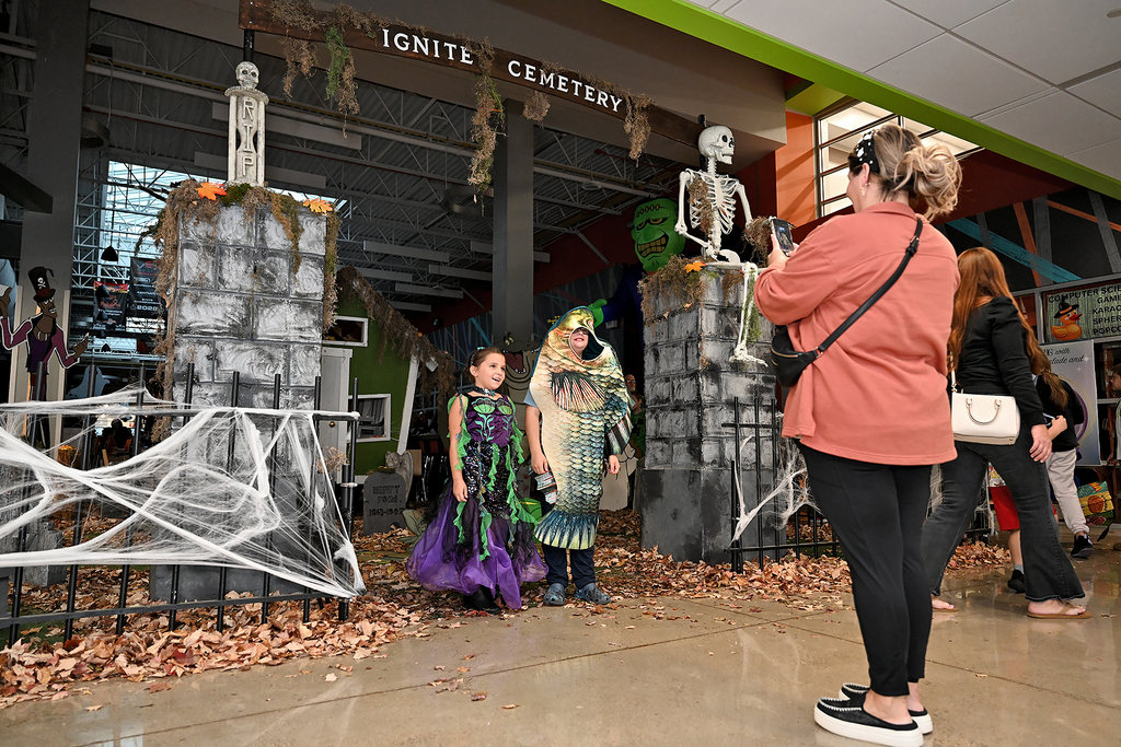 Two young girls in Halloween costumes stand by a skeleton gate. A woman in a pink coat snaps a photo.