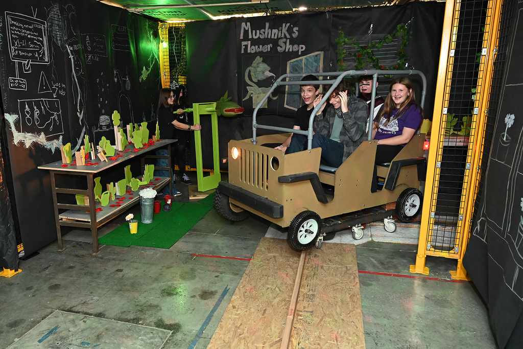 Four students sit inside a small, boxy vehicle with green paint in a room. A table holds objects.