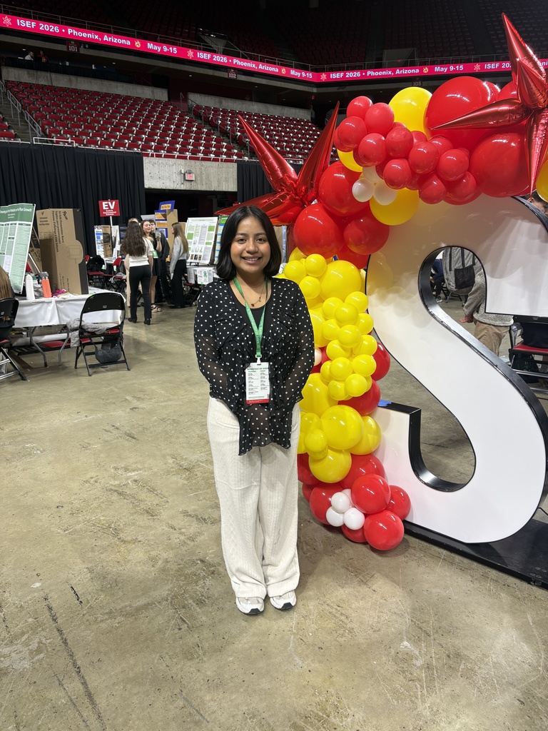 Person standing indoors next to a large white letter 'S' decorated with red, yellow, and white balloons at an event venue.