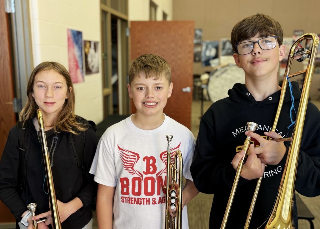 Three young students holding brass instruments, standing in a school music room with instruments and posters in the background.