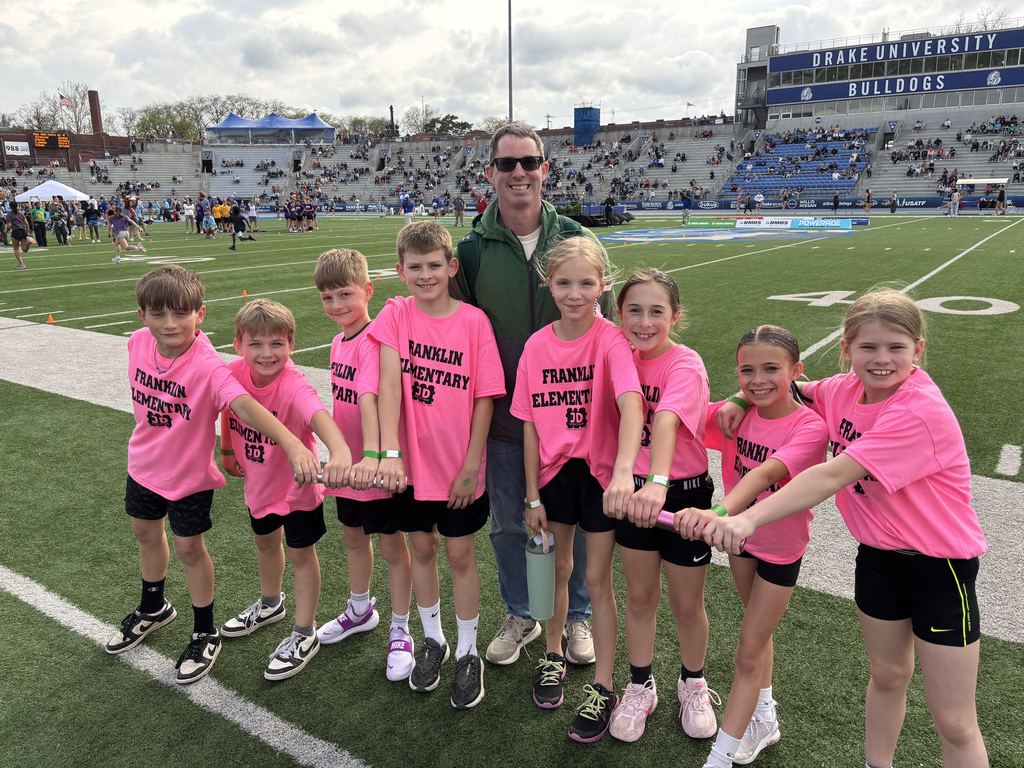Eight students standing on a football field in a line and holding a track baton, with their coach standing behind them.