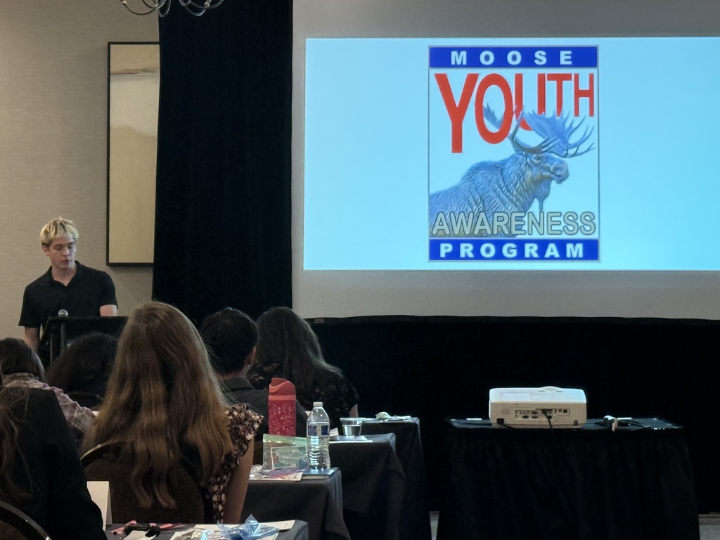 Audience attending a presentation on the Moose Youth Awareness Program with a speaker at the podium and a projected program logo.