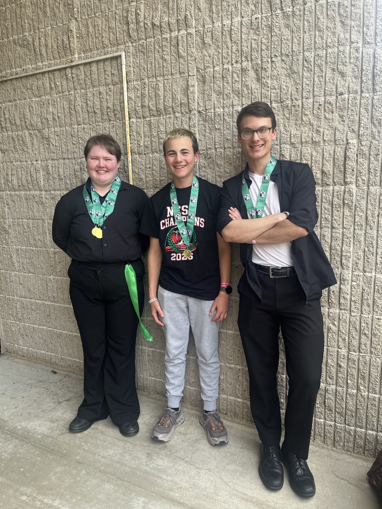 Three young men standing against a beige brick wall, each wearing medals around their necks, celebrating an achievement.