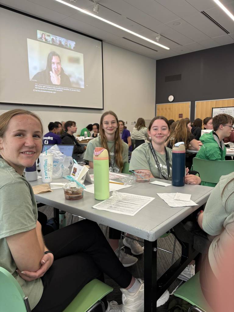 Three students smile at a table during a school literacy event as a presentation plays on a large screen behind them.